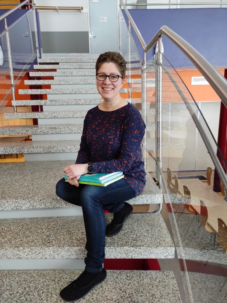 Kuvassa erityisopettaja Hanne Bräysy istuu yläkoulun toiseen kerrokseen vievillä portailla. Hän hymyilee. Hänellä on sylissä kirjoja. 